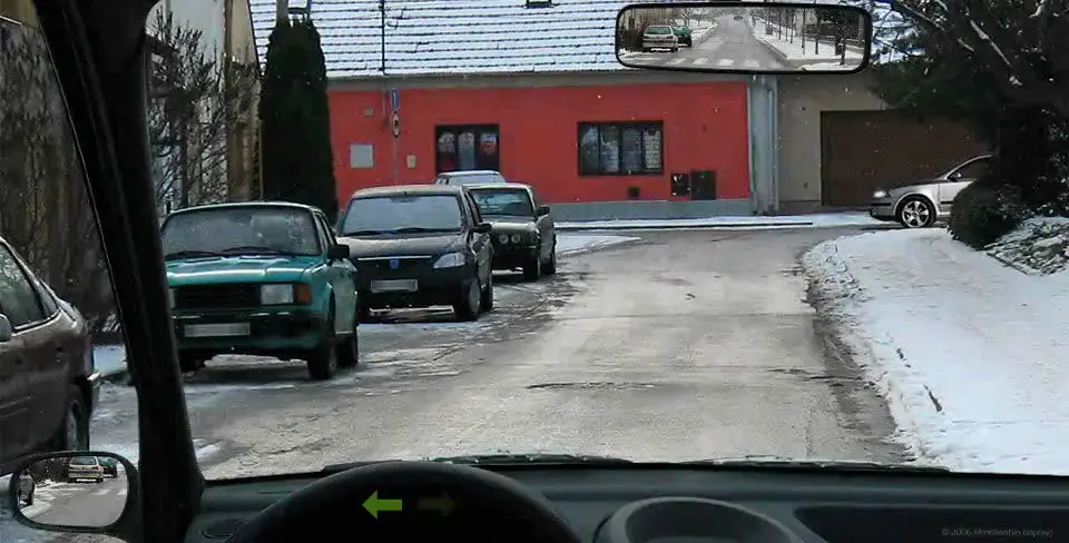 Pohled z auta s rozsvíceným levým blinkrem na úzkou ulici se zaparkovanými auty vlevo a stříbrným autem vpravo