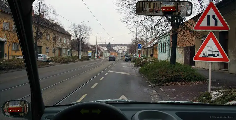 Pohled z auta na ulici s tramvajovými kolejemi vlevo a výstražnými značkami Zúžená vozovka a Tramvaj vpravo