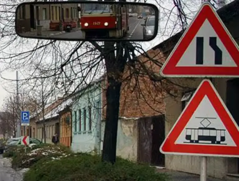 Detail zpětného zrcátka, ve kterém je vidět červená tramvaj, a pod ním dvě výstražné značky
